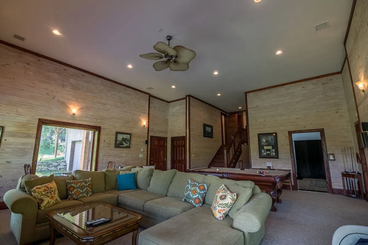 A cozy lower-level space with a large sectional sofa, a wooden coffee table, and a pool table. The arrangement encourages both competitive play and communal lounging, with the staircase and open layout hinting at easy access and room for gatherings.
