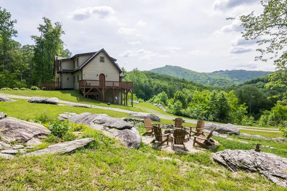 The image presents the house and its large fire pit set amidst a sweeping mountain landscape. Guests are shown gathered around the fire, enjoying the warmth and camaraderie, with trails and greenery inviting exploration beyond the yard.