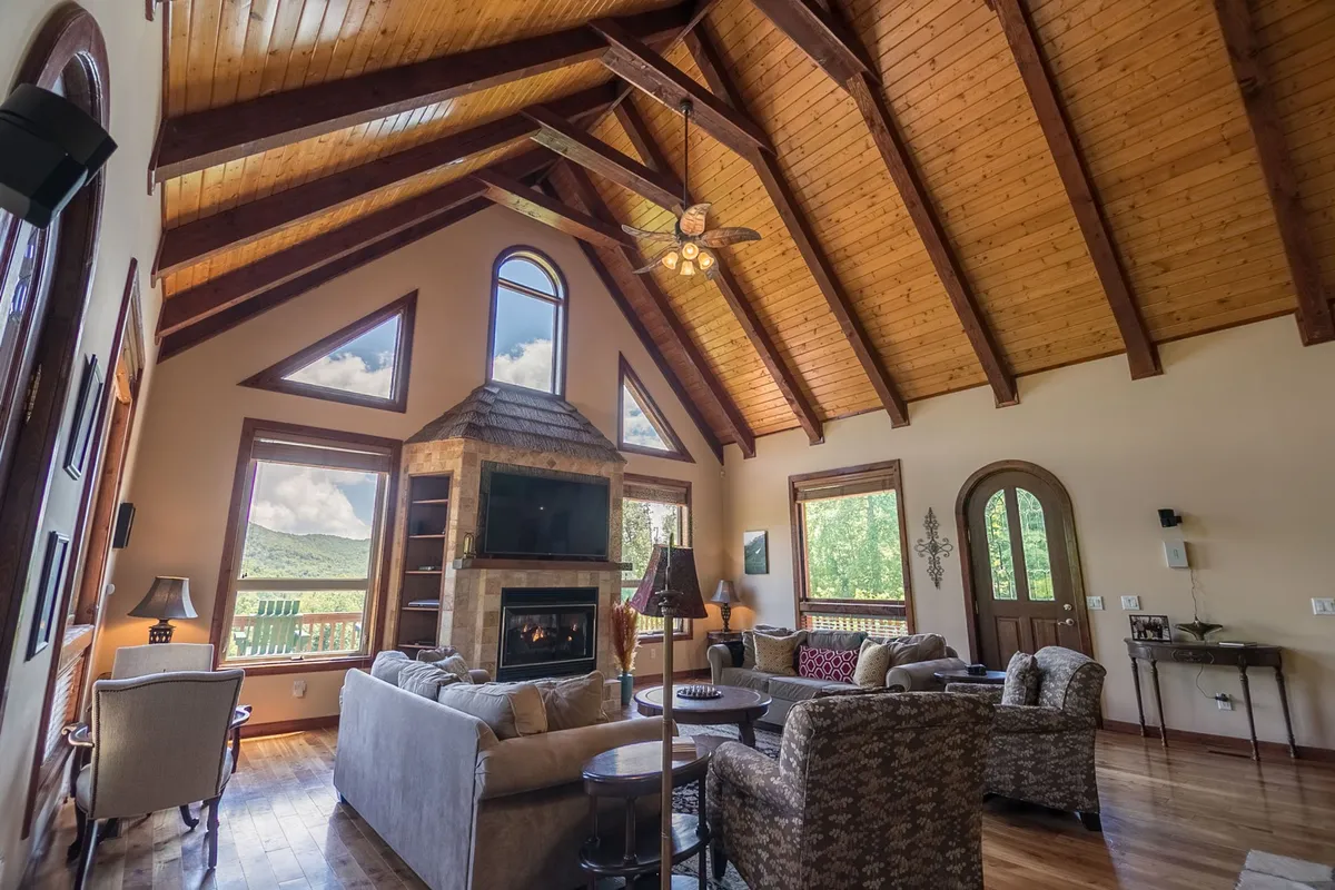 A cozy, inviting living space centered around a prominent stone fireplace. Large windows let in ample light, while a sofa set and wooden beams create a welcoming atmosphere for relaxation or socializing. The fireplace acts as a natural focal point for groups to gather, echoing the section's mention of a three-sided stone fireplace.