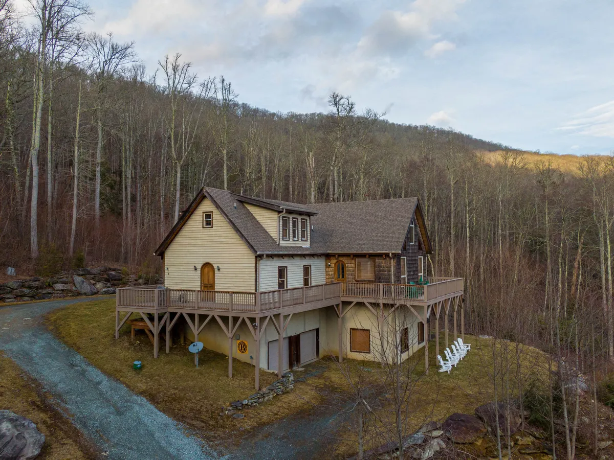 The property's structure is framed by trees, with a pathway leading to the house and a generous outdoor deck. The setting appears peaceful and private, with plenty of open, grassy areas for pets to roam.