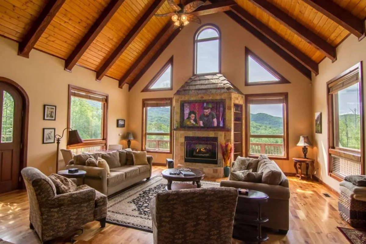 The image showcases a cozy, stylish living area with plush seating arranged around a striking central fireplace. Expansive windows reveal breathtaking mountain and sunset views, flooding the room with natural light and offering a seamless connection to the stunning outdoors. The scene invites relaxation, conversation, and shared moments with family or friends in a serene, private mountain retreat.