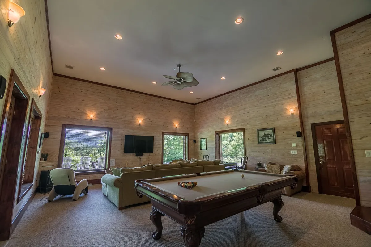 A lively game/media room featuring a pool table as the centerpiece, surrounded by comfortable seating and large windows that flood the space with natural light. A TV is mounted on the wall, and the casual arrangement invites guests to gather for game nights, movie marathons, or relaxed socializing.