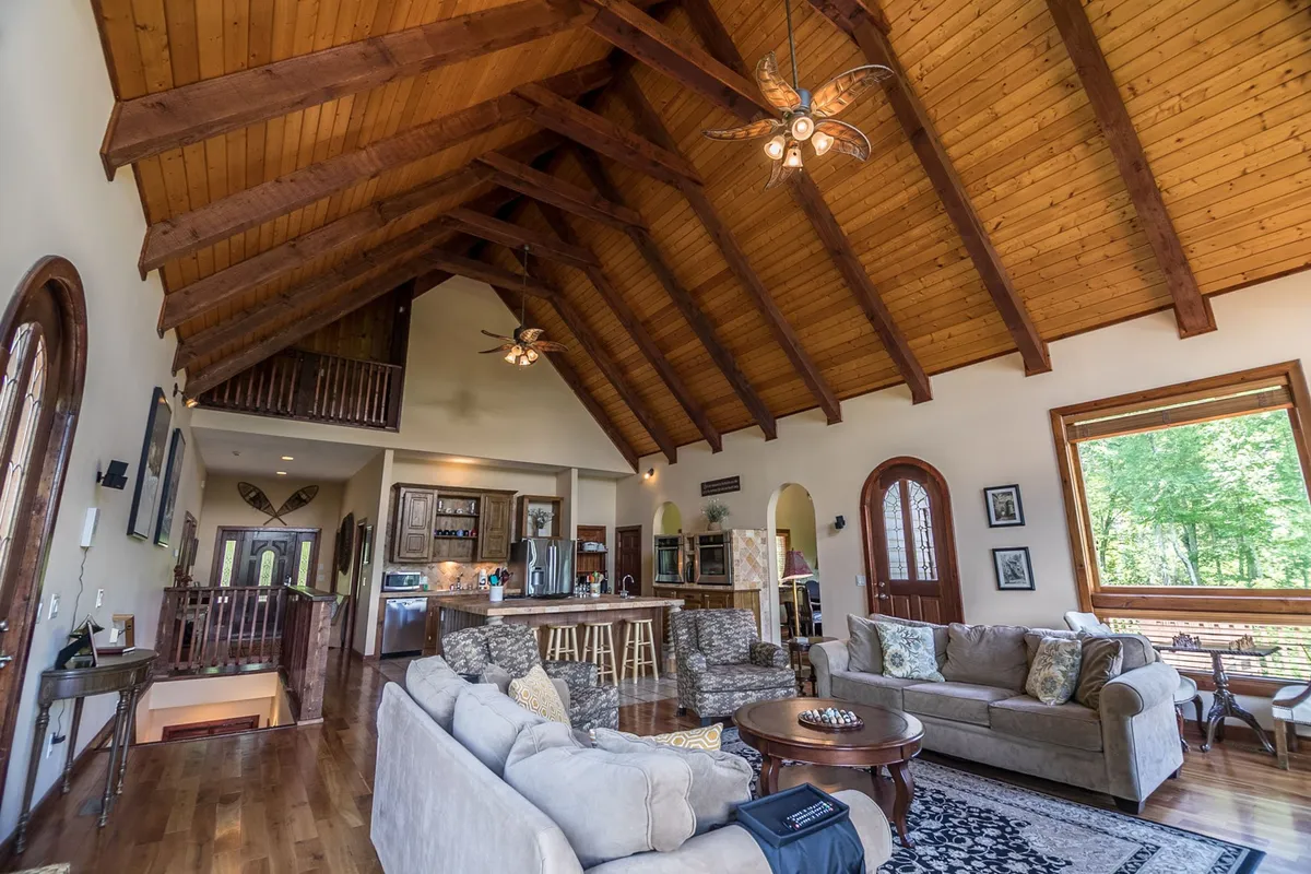 A spacious great room with a high wooden ceiling and large windows allowing natural light to flood the comfortable seating area. The kitchen island is visible in the background, seamlessly connecting the lounge area to the kitchen. The arrangement is inviting, warm, and designed for gatherings, with plush sofas and a view toward the heart of the home.
