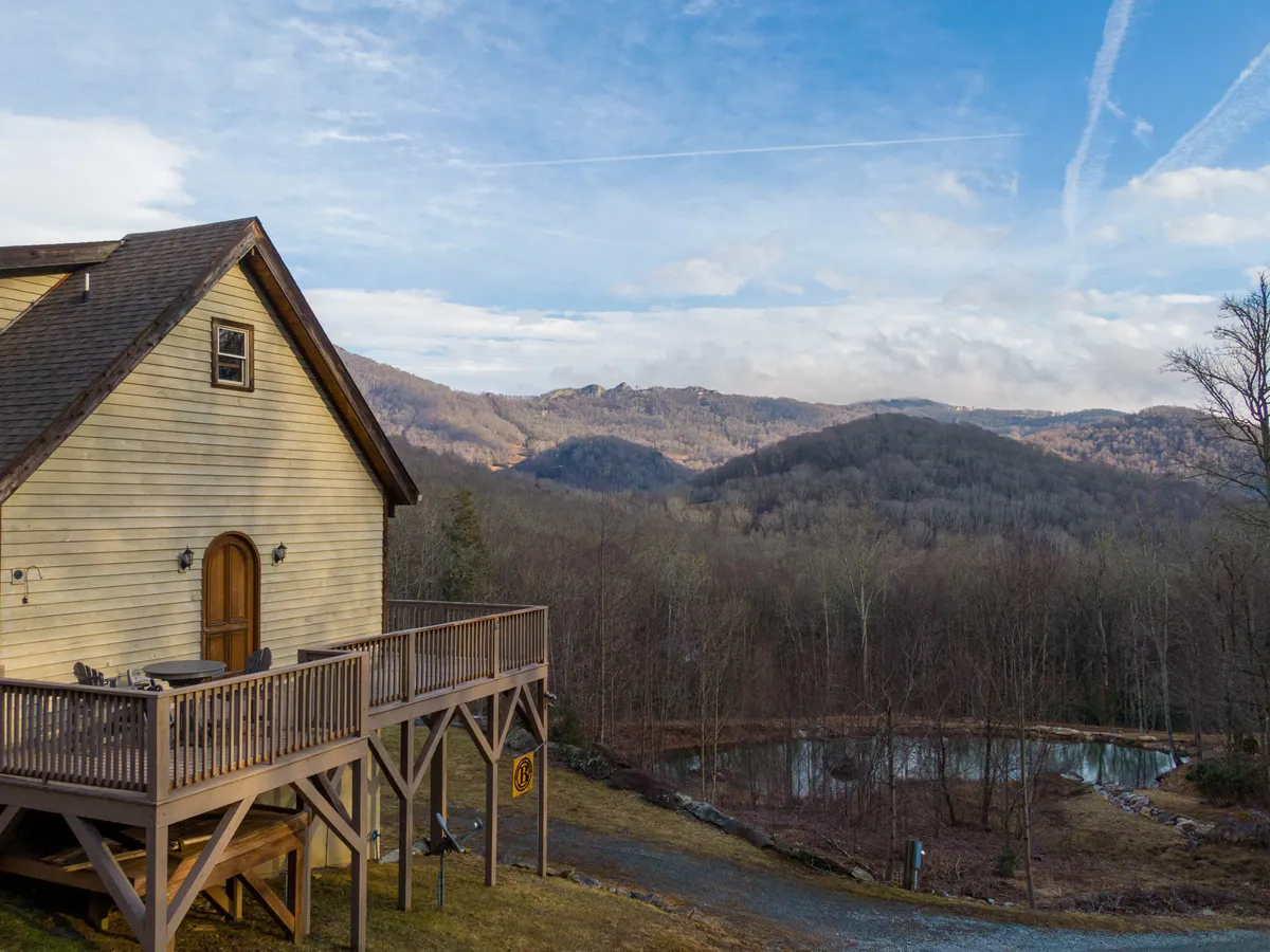 The image captures the manor's deck overlooking rolling mountains and a tranquil pond, surrounded by trees. The scene invites exploration, with clear sightlines into the natural landscape, suggesting opportunities for hiking and wildlife watching directly from the property.