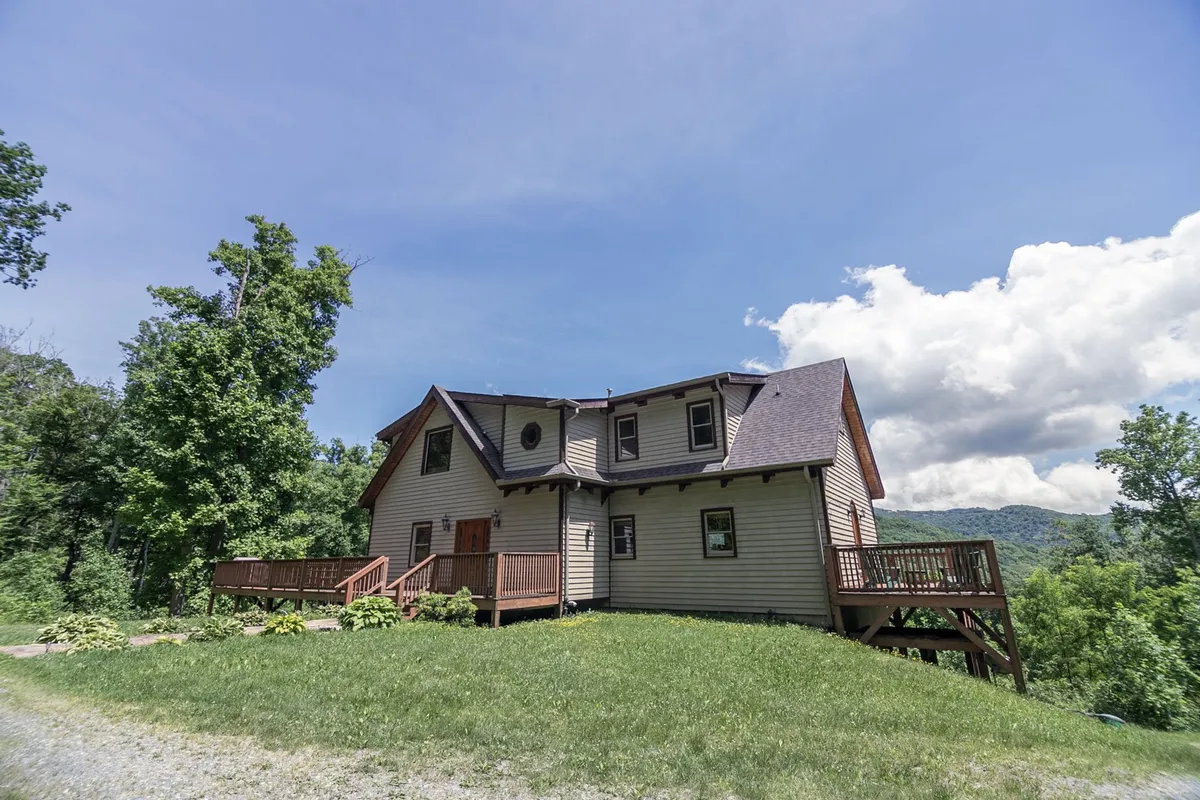 A spacious wooden deck extends from the house, surrounded by lush greenery. The unique roofline and natural landscape create an inviting atmosphere for gatherings, and there is ample open space for both people and pets to relax or play. The setting suggests easy access to the outdoors, perfect for families and their furry companions.