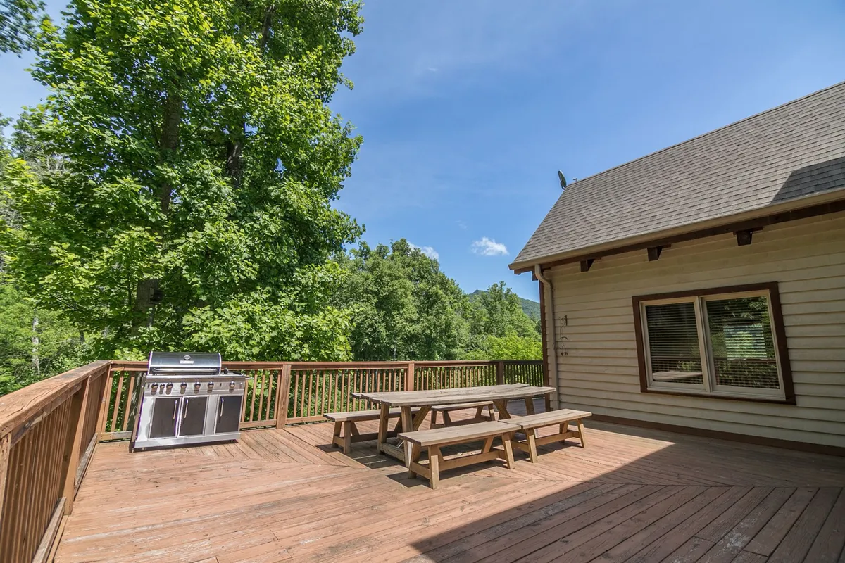 An inviting exterior deck with a grill, table, and chairs surrounded by lush trees. The deck is designed for gatherings, with ample seating for outdoor dining, socializing, and enjoying the natural surroundings. The house exterior is visible, linking the outdoor space to the main living areas inside.