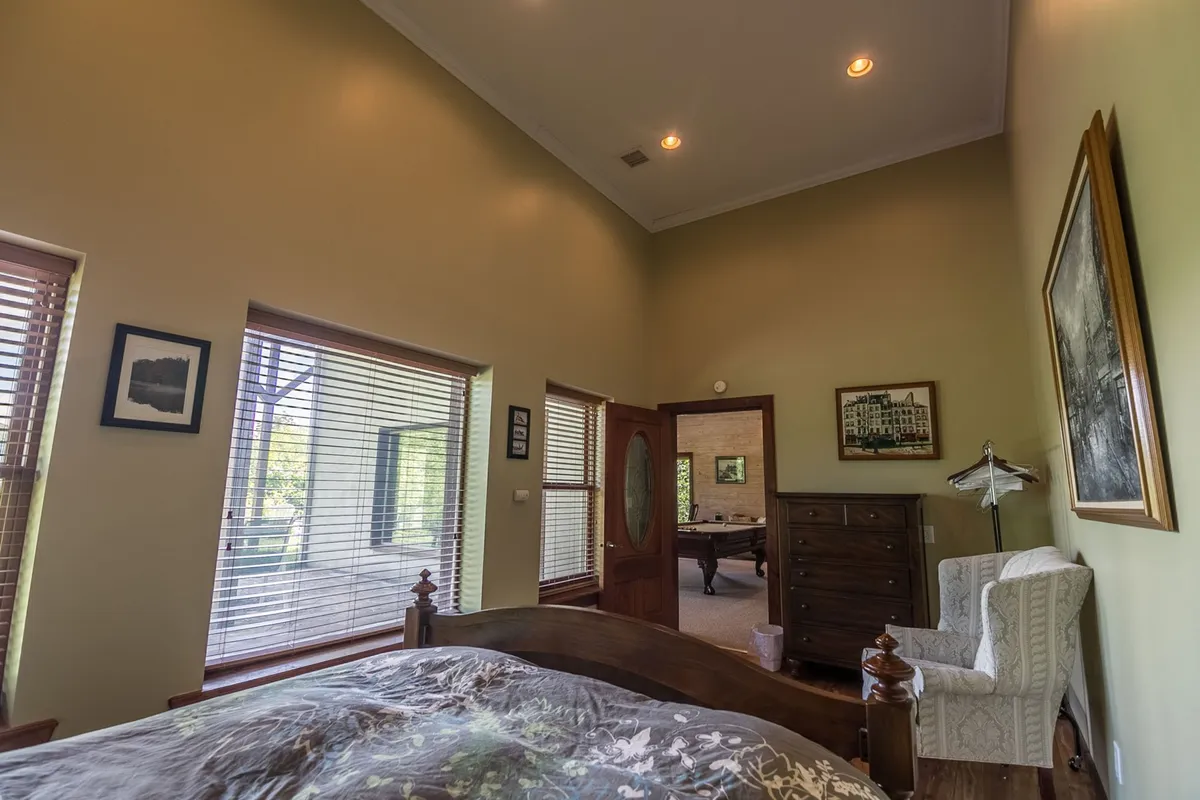 A cozy bedroom with intricate bedding on the bed, large windows with adjustable blinds, a wooden dresser, a comfortable armchair, and curated wall art. The room is perfect for unwinding, enjoying a morning coffee, or reading by the window.