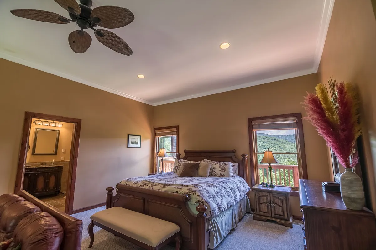 A spacious bedroom featuring a king-sized bed positioned to face large windows that frame a mountain view. The room is styled with wooden furniture, soft decorative plants, and warm, calming tones. Gentle natural light filters in, lending the space a serene and luxurious feel.